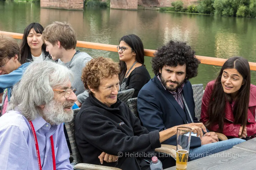 A white man with shaggy white hair and a beard sits next to an older white woman with brown hair and a younger man with dark hair and a beard. A young woman with long dark hair is to the far right.