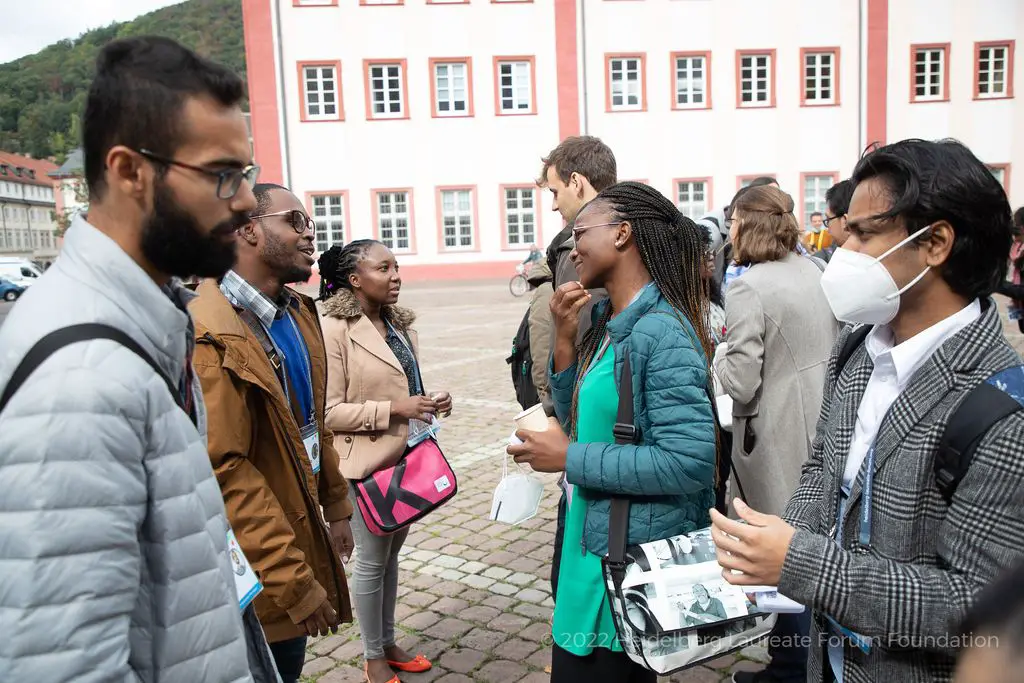A group of people of different races in a cobbled square in Heidelberg. One of the conversants is wearing a white N-95 mask.